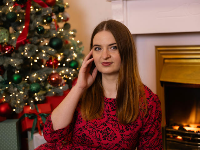 FreyaDak with straight brown hair, blue eyes, wearing a red and black floral dress, touching her cheek, standing in front of a lit fireplace and decorated Christmas tree.