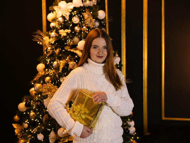 FreyaDak with brown hair, white turtleneck sweater, holding gold gift, standing in front of a decorated Christmas tree. She's got a cute smile and a curvy ass.