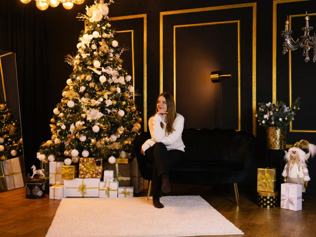 FreyaDak with long brown hair, white sweater, black pants, and black socks, sitting on black velvet sofa, Christmas tree with gold and white ornaments, and wrapped gifts nearby.