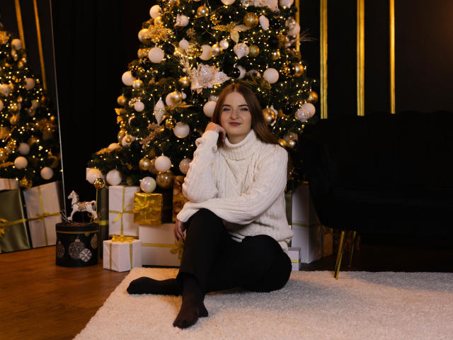 FreyaDak with brown hair, white turtleneck, black pants, and socks, sitting on a white rug, hand on chin, in front of a decorated Christmas tree with gold ornaments.