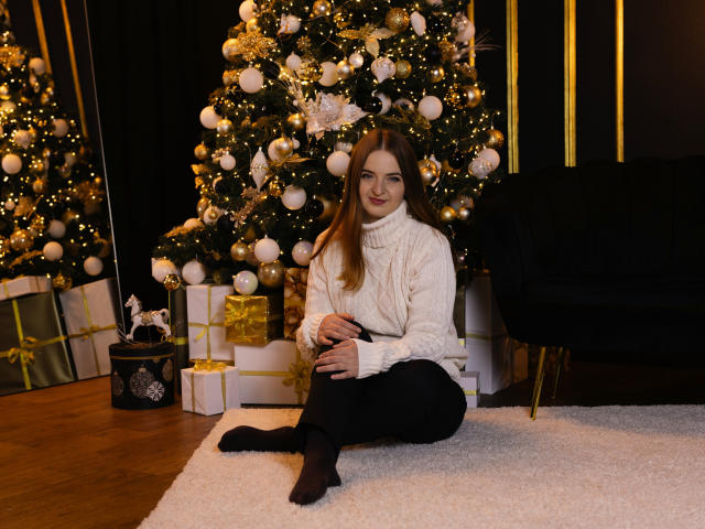 FreyaDak, sitting on a white rug, wearing a white turtleneck and black pants, with brown hair, smiling, in front of a decorated Christmas tree, black chair to the right.