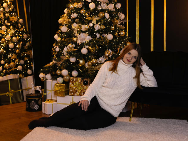 FreyaDak, with long brown hair, white turtleneck, and black pants, leans on a black chair, legs out, next to decorated Christmas trees and gift boxes, smiling at the camera.