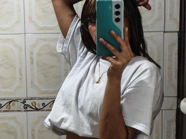 AmyLennox with medium tits, wearing a white tee, holding a teal phone, raising her arm, showing a tattoo on her upper arm, in a tiled bathroom.