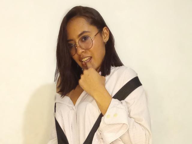 AmyLennox with dark brown shoulder-length hair, wearing glasses, a white striped shirt, and black suspenders, biting her finger with a playful expression.