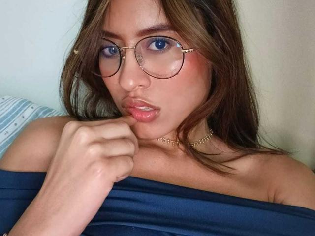 AmyLennox has light brown skin and long brown hair. She wears round, gold-rimmed glasses with blue lenses and a gold chain necklace. Her lips are slightly parted with a subtle pinkish-red lipstick. She holds a dark blue off-shoulder garment with her right hand, her fingers gently pressing it against her collarbone. Her expression is neutral, with a slight hint of curiosity. The image is a close-up portrait. The overall composition focuses on AmyLennox's face and upper chest.