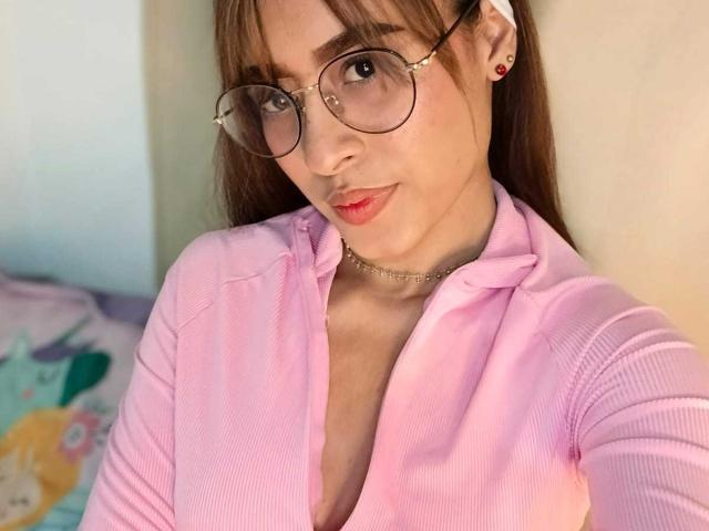 AmyLennox with glasses, pink ribbed shirt, small tits, light skin, red earrings, choker, subtle red lipstick, brown hair, slight smile, soft lighting, bedroom setting, selfie.