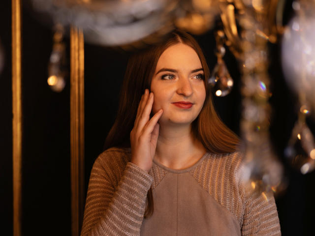 FreyaDak with long brown hair, hand on cheek, smiling, wearing a beige sweater with textured sleeves, looking to the side, soft candlelight reflecting in her eyes.