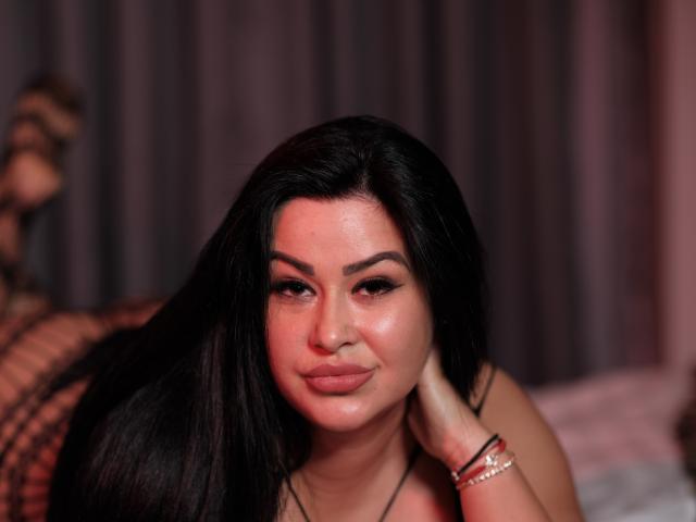 AilynDiamond with long black hair, pouty pink lips, and a slight smile, resting her head on her hand, wearing a bracelet, looking seductive.
