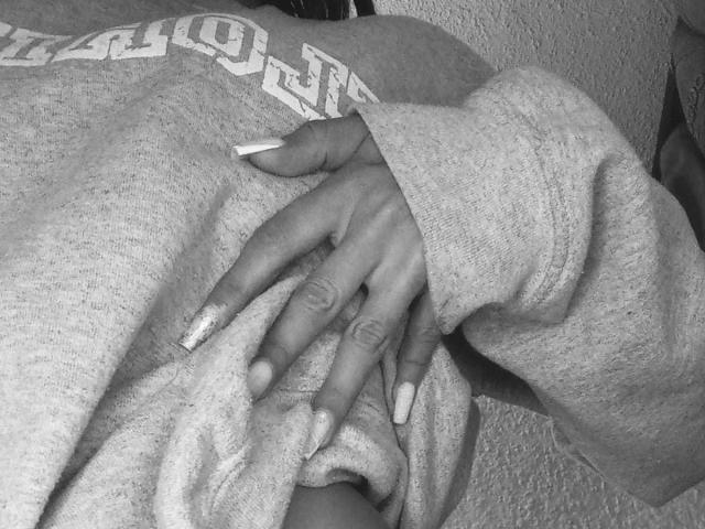 Black and white photo of AmyLennox's hand resting on their chest. AmyLennox's hand has long, white-painted nails with a glittery texture. They wear a loose, textured, gray sweater with a visible "UNO" logo. The sweater's fabric is slightly wrinkled. AmyLennox's skin is dark, and their fingers are slightly curled. The texture of the sweater and the glittery nails are the main focal points. The image has a close-up, intimate composition.