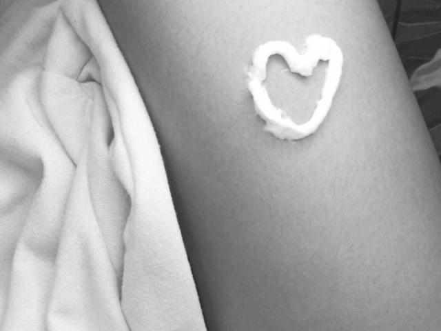 Black-and-white close-up of AmyLennox's arm. A small, heart-shaped smear of white cream or lotion sits on the skin. The arm is slightly angled, with a soft, textured cloth partially covering the left side. The cream is slightly uneven, with rough edges. Skin is smooth and light in tone. Simple composition with focus on the heart shape and cream texture. Slight shadow on the arm adds depth. AmyLennox's arm is in the center, cream is to the right.