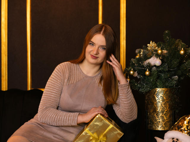 FreyaDak with red hair, wearing a beige dress, holding a gold-wrapped box, sitting next to a decorated Christmas tree, smiling, touching her hair.