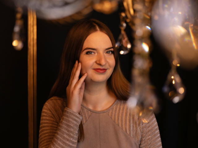 FreyaDak with long brown hair, fair skin, and light makeup, wearing a sparkly beige top, touching her cheek with a gentle smile, surrounded by glowing, ornate chandelier.