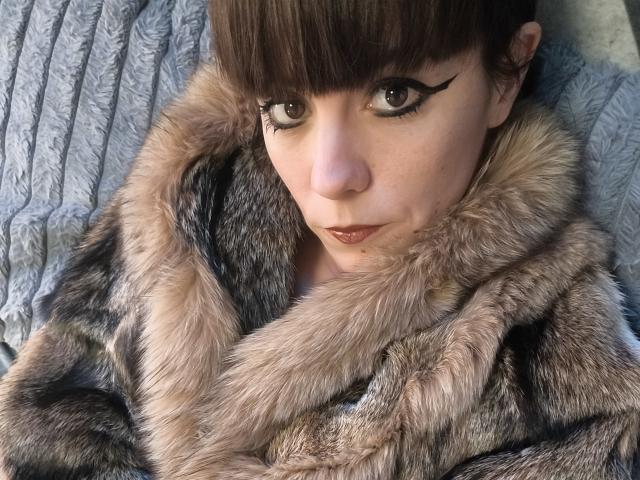 OrianaLaFrancaise, con i capelli castani, la frangia e l'eyeliner marcato, indossa un cappotto di pelliccia grigia e guarda la fotocamera con un'espressione sottile e leggermente accigliata..