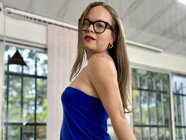 PamelaColin with glasses, blue dress, and earrings, standing with a smile, showing her tits and freckled shoulders, in a bright room with windows.