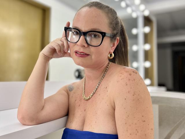 PamelaColin with glasses, blue strapless top, gold earrings, necklace, freckled shoulders, and brown ponytail, leaning on white table, smiling, hand near face.