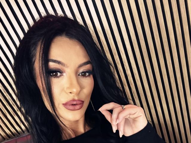 KinkyAmelie with black hair, full lips, and dramatic makeup, holding her ringed finger, wearing a black top, looking at the camera with a slight pout.