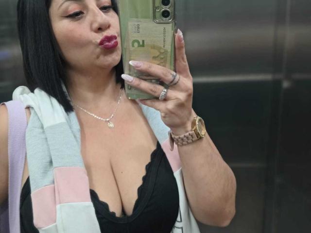 AngeleRoux met volle, roze lippen, een zwarte kanten top, grote borsten, die een telefoon vasthoudt, draagt een roze en witte gestreepte vest, een zilveren ketting en een horloge, in een lift..