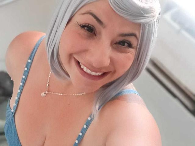 AngeleRoux, com um corte de cabelo curto prateado, sorrindo, vestindo um vestido azul com bolinhas brancas, mostrando o decote e usando um colar de ouro. Seus seios estão ligeiramente visíveis..