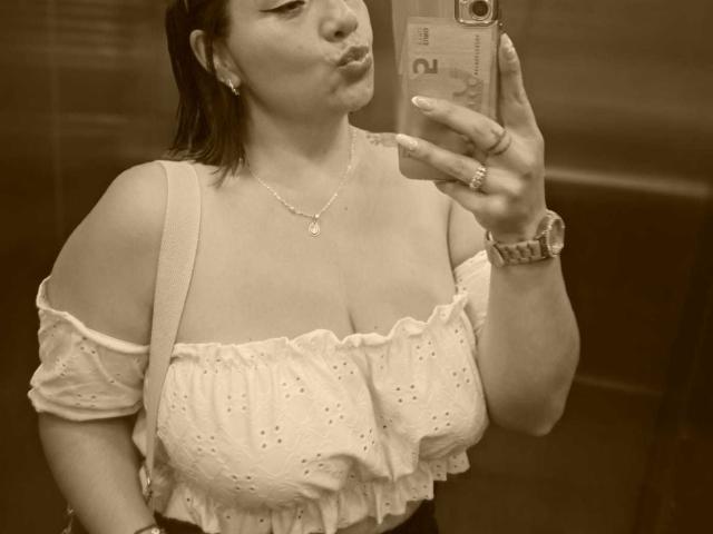 AngeleRoux taking a mirror selfie, pouting lips, wearing a white off-shoulder top with small holes, showing her big tits, holding a phone with a ring on her finger.
