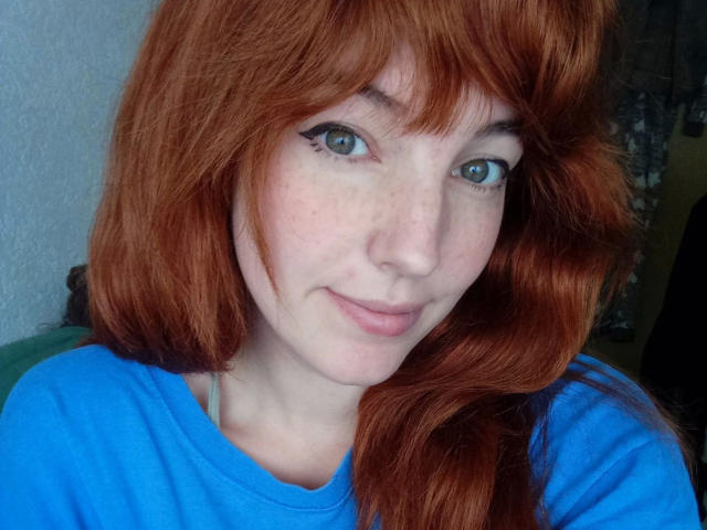 LoveKiitsune-hot with red hair, blue eyes, and freckles wearing a blue shirt, smiling slightly, has a cute face and wavy hair, looking directly at the camera.