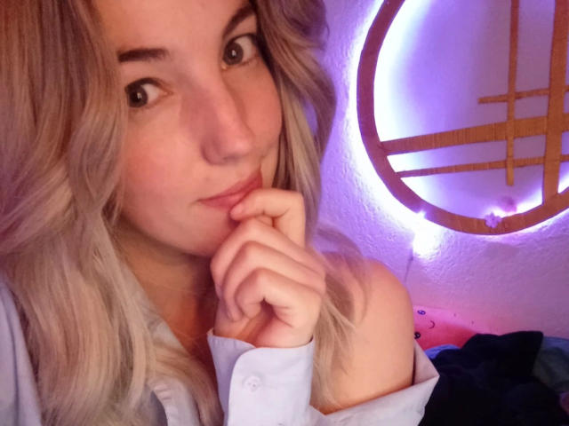 LoveKiitsune-hot with blonde hair, looking at camera, thinking, hand near mouth, wearing light shirt, purple neon ring light in background, soft lighting, cute, slightly pouty expression.
