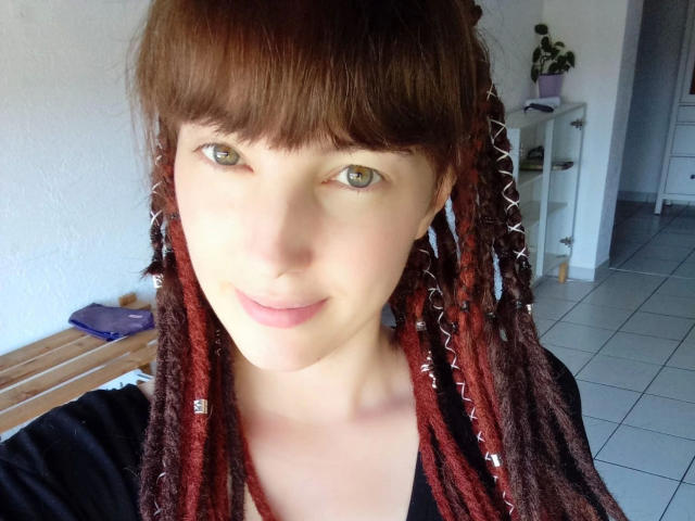 LoveKiitsune-hot has red-brown dreads, fair skin, green eyes, and a slight smile, wearing a black shirt, with a wooden bench and white tiled floor in the background.