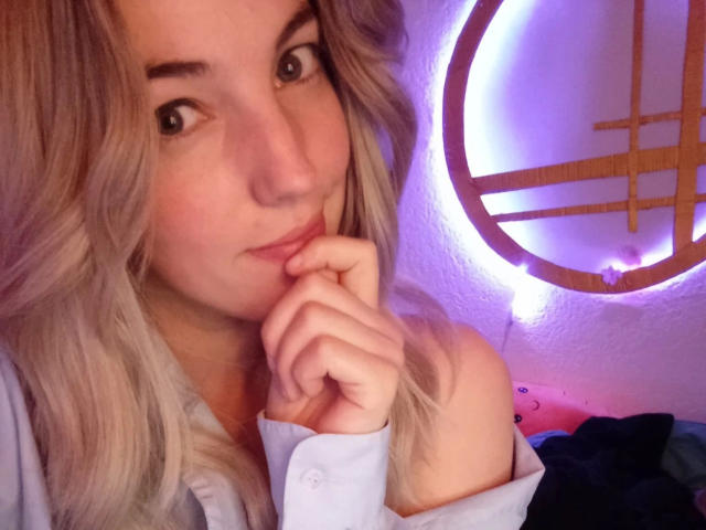 LoveKiitsune-hot with blonde hair, looking cute and thinking, has her hand near her mouth, wearing a light blue shirt, under purple neon light.