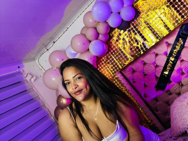 DannaRamiirez with black hair, medium tits, and pink makeup, smiling in a room with purple and pink balloons, and a gold sequin wall. She's wearing a white tank top.