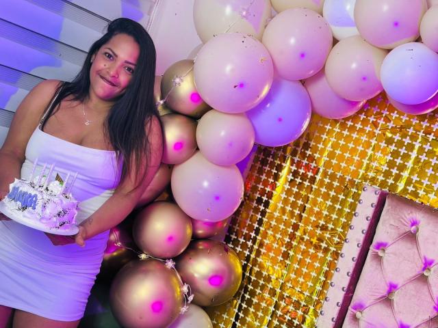 Danna Ramirez, con lunghi capelli neri, indossa un abito bianco, tiene in mano una torta con decorazioni bianche e blu, ed è circondata da palloncini bianchi e dorati, sorridendo alla macchina fotografica..