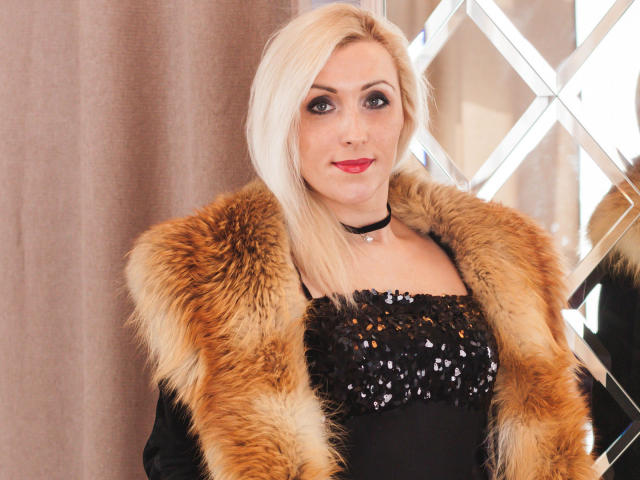 MistyLady with blonde hair, red lipstick, and black sequin dress, wearing a furry brown coat, standing in front of mirrored wall, looking at camera with a slight smile.