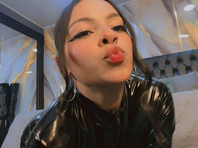 LindaFrancoo with pouty red lips, black winged eyeliner, and glossy black latex outfit, wearing large hoop earrings, in a bedroom with marble-patterned walls.