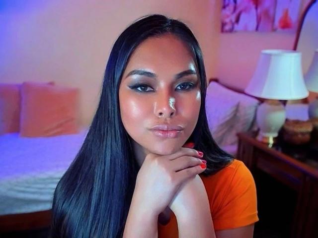TsLexie-hot with shiny black hair, orange top, and red nails, resting her chin on her hands, giving a smoky-eyed, glossy-lip look, in a softly lit bedroom.