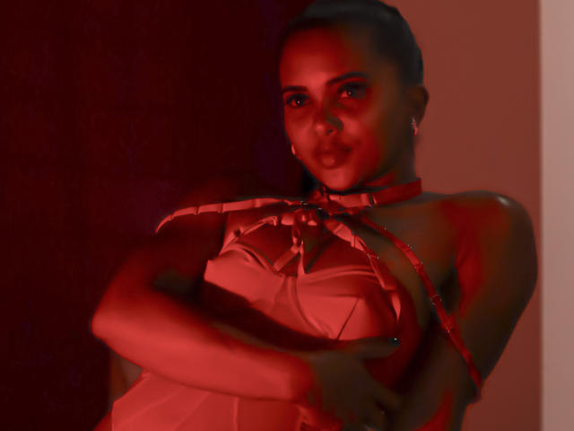 ZaraSolei in red lingerie, covering her tits with arms, wearing a choker, red light casting shadows on her face, dark hair in a bun, subtle smile.