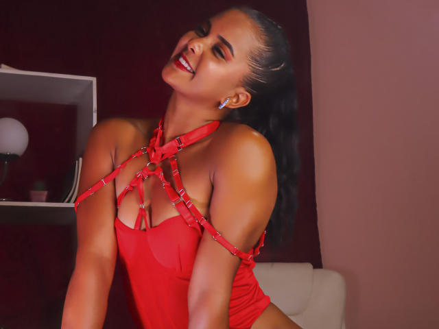 ZaraSolei with dark hair in a ponytail, smiling, wearing a red harness that exposes her tits and red mesh top, leaning forward, showing her ass slightly.