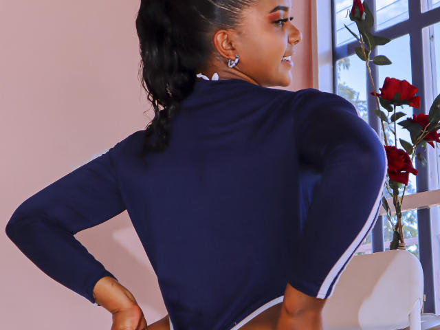 ZaraSolei's back, showing a tight navy blue top, with hands on her hips, black braided ponytail, and red rose earrings. She's smiling, looking over her shoulder.
