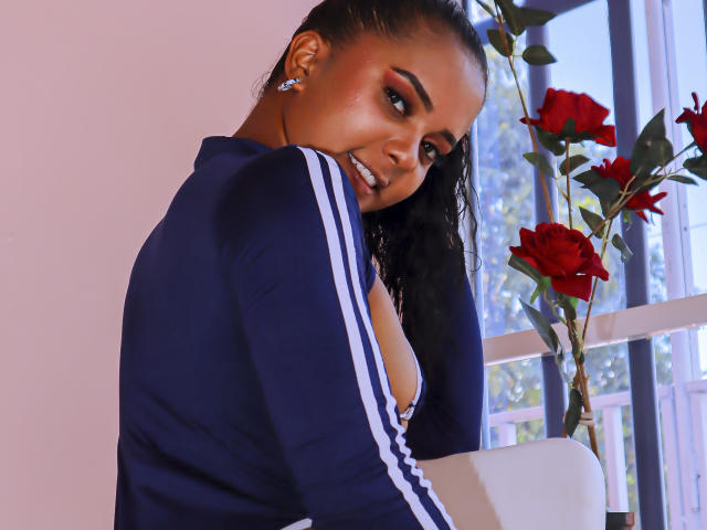 ZaraSolei with dark skin, curly black hair, wearing a blue track jacket, showing her tits, smiling, red roses in background, earrings, natural light, window.
