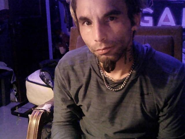 RibBlair with a goatee, wearing a dark grey shirt and gold chain, has a serious expression, sitting in dim blue lighting, with a brown chair behind him.