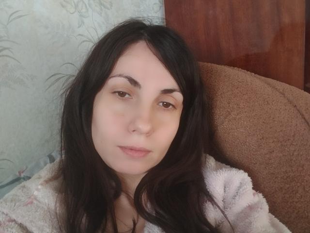 AvaBobby has pale skin, dark brown wavy hair, and brown eyes. She's wearing a pink fuzzy sweater, sitting in a brown chair with a wooden door behind.