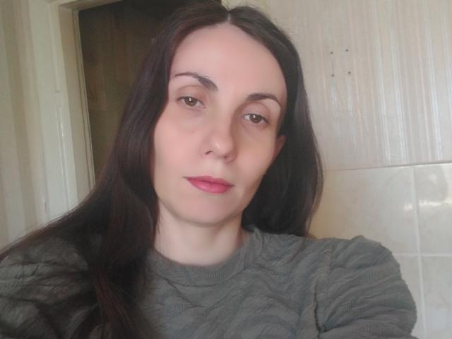 AvaBobby has long dark brown hair, pale skin, brown eyes, pink lipstick, and wears a gray sweater. Her tits are small, and she's giving a slight, confident smirk.