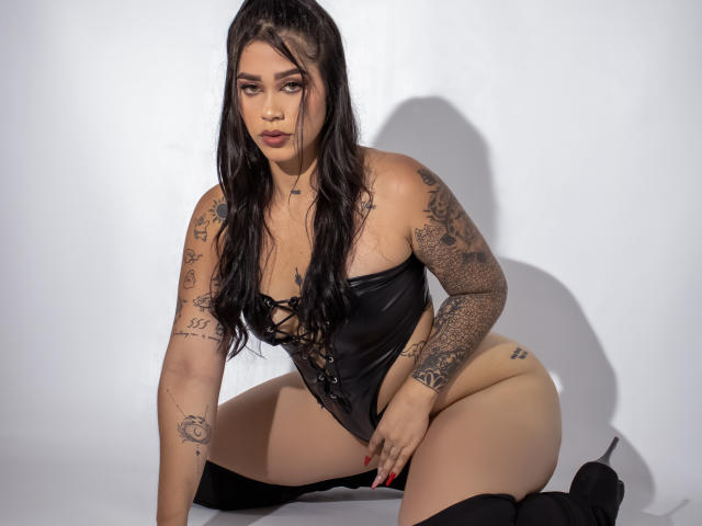 CelesteSwith, tattooed with tattoos on arms and thighs, kneels in black leather corset, lace-up front, and thigh-high boots, showing her big ass and small tits, with long black hair.