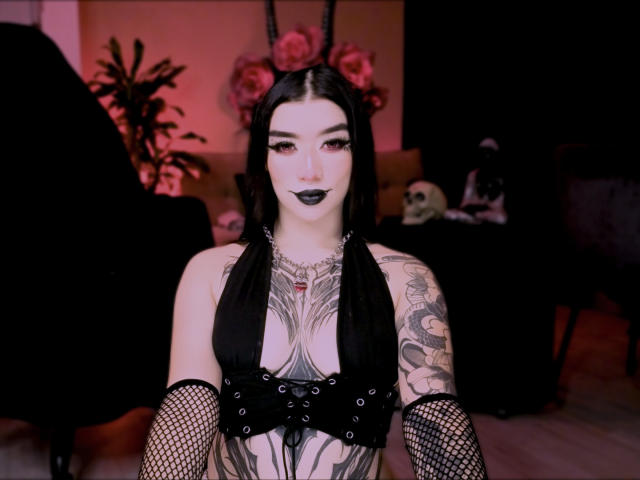 VanessaBlackRose with black hair, tattoos, black lipstick, black lingerie, fishnet arms, and visible tits, standing in a goth-themed room. Her tattoos include a large chest piece.