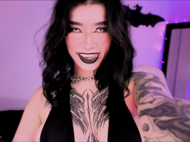 VanessaBlackRose with black lipstick, tattoos on chest and arm, chain necklace, black vest, and wavy dark hair, smiling with her tongue out, purple light in the background.