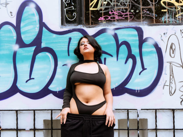MisakiWong with black hair, wearing a cutout black top and black pants, standing in front of graffiti, showing her tits and a bit of her pussy through the top.