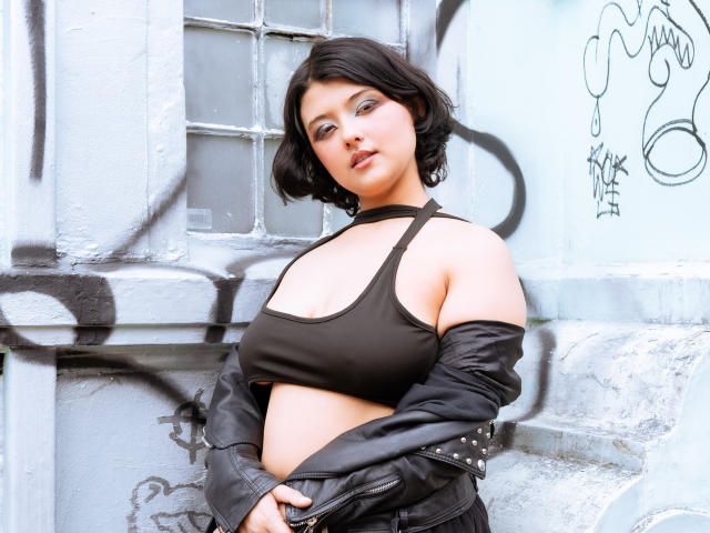 MisakiWong with short black hair, wearing a black halter top and leather jacket, showing her tits and belly button, standing against a graffiti-covered wall.