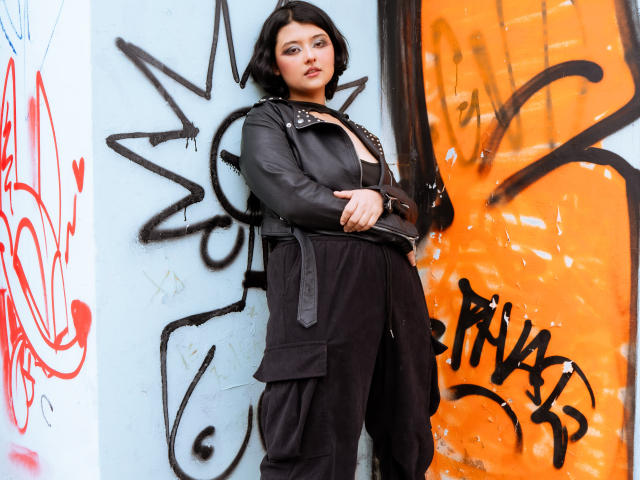 MisakiWong with short black hair, wearing a black leather jacket and black pants, standing against a graffiti-covered wall, hands in front, looking confident, with a subtle smile.