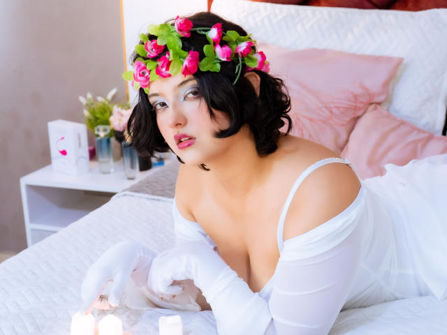 MisakiWong with short black hair, pink rose and green leaf headpiece, white off-shoulder dress, white gloves, lying on bed, looking up, pink pillow, white bed, candles.