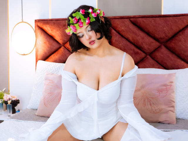 MisakiWong with flower crown, white off-shoulder shirt, and large tits, sitting on bed, looking down, pink floral pillows, red velvet headboard, hanging light, soft natural light.