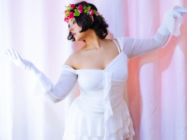 MisakiWong with short black hair, white off-shoulder dress, white gloves, and pink flower headpiece, showing off her tits and curvy ass, arms raised, smiling.