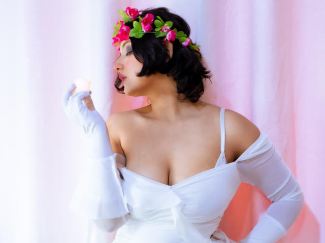 MisakiWong with short black hair, white off-shoulder dress, and white gloves, wearing a pink and green flower crown, touching her lips, showing large, round, perky tits.