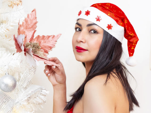 ChanelBrouw with dark hair, red lipstick, and Santa hat, touching pink poinsettia on white Christmas tree, showing bare shoulders, red dress, and white background.